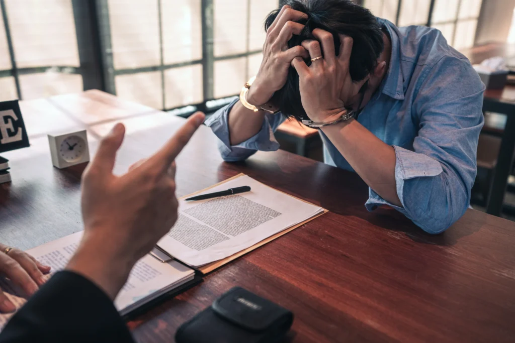 A person in handcuffs, sitting at a desk, holding their head.
