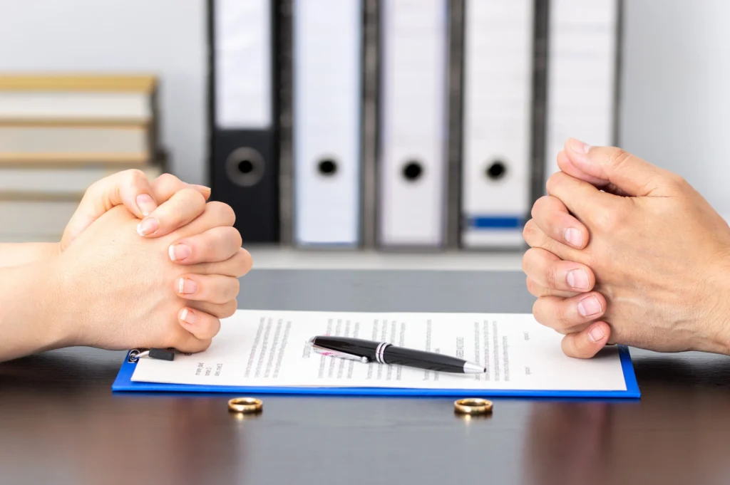Two people with their hands crossed on top of a clipboard with paperwork and a pen and two golden rings on the table.
